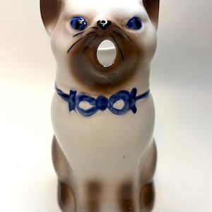 May include: A ceramic Siamese cat figurine with a cream-colored body, brown ears, face, and paws, featuring bright blue eyes and a painted blue bow tie around its neck. The cat has an open mouth, creating a hollow opening in the center of its face.