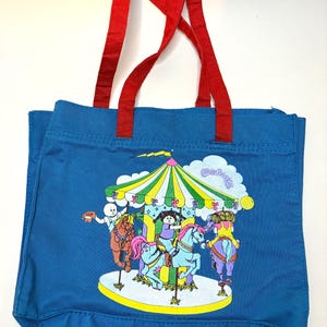 May include: A blue canvas tote bag with two red fabric handles. The front features a colorful graphic print of a carousel with Cabbage Patch Kids characters riding horses, under a green and yellow striped canopy, with a white cloud in the background.