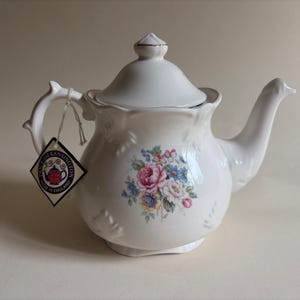 Op de afbeelding: Een witte keramische theepot met een bloemenboeketpatroon in roze, blauwe en gele tinten. De theepot heeft een geschulpte rand, een deksel met een goudkleurige knop en een ruitvormig label met de tekst Price and Kensington Made in England.