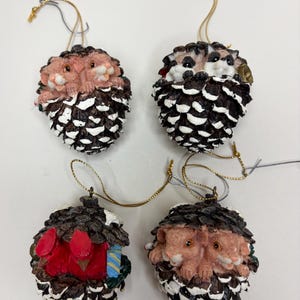 May include: Four decorative ornaments shaped like pinecones with white-tipped scales. Three ornaments feature pairs of small tan animals with dark eyes, while one ornament features two bright red birds with a small blue and yellow gift box.