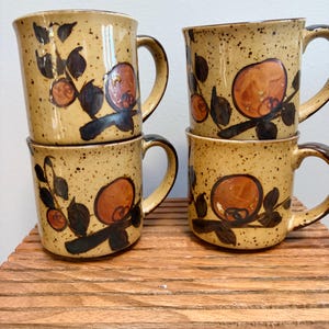 May include: A set of four ceramic mugs with a speckled tan glaze, each featuring a hand-painted design of dark brown branches and orange fruit. The mugs have a cylindrical shape with a curved handle on the side and a smooth finish.