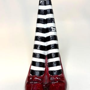 May include: A decorative figurine featuring two legs wearing black and white horizontally striped stockings, ending in a pair of shiny, glittery red shoes with small bows on the front. The legs are positioned vertically, joined together at the top.