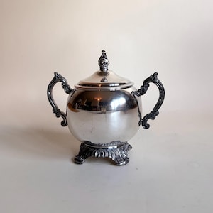 Op de afbeelding: Een gepolijste zilverkleurige suikerpot met bijpassend deksel, voorzien van twee sierlijke, gebogen handvatten en een decoratieve voet met gedetailleerd krulwerk. De pot heeft een glad, rond lichaam en een deksel met een kleine knop.