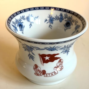 May include: A white porcelain bowl featuring a blue floral garland pattern around the rim and interior. The exterior displays a red flag with a white star above a banner with the text WHITE STAR LINE in red lettering and a dark blue border.
