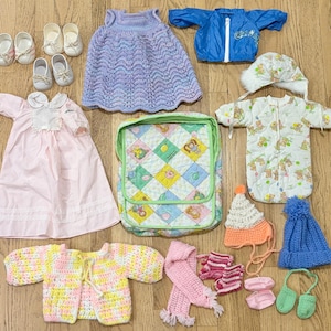 Puede incluir: Una colección de ropa y accesorios para muñecas que incluye un vestido de punto lavanda, un cortavientos azul, un vestido rosa, un bolso acolchado con patrón de diamantes, un traje de nieve blanco estampado con capucha, un cárdigan multicolor, gorros, bufandas y cuatro pares de zapatos.