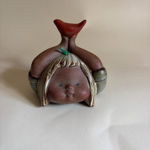 Może przedstawiać: Ceramiczna figurka przedstawiająca zaokrągloną twarz z małymi niebieskimi oczami i ustami, zwieńczona czerwonym elementem w kształcie ptaka. Postać ma jasne włosy z zielonym akcentem liścia oraz brązowe, nakrapiane, szkliwione wykończenie.