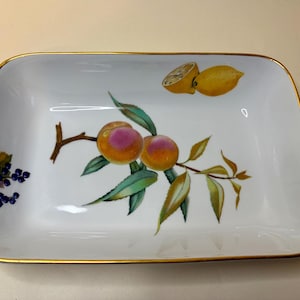 Può includere: Un vassoio rettangolare in porcellana bianca con bordo dorato. Il centro presenta un'illustrazione botanica di due pesche su un ramo con foglie, un grappolo di bacche blu scuro a sinistra e un limone giallo nell'angolo in alto a destra.