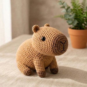 May include: A small, tan, crocheted capybara figurine with a rounded body, short legs, and dark brown accents on the feet and snout. The toy features a textured yarn surface, small round black eyes, and a stitched nose detail on its face.