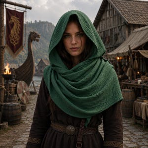 May include: A textured forest green hooded cowl made of lightweight fabric draped over the head and shoulders, worn over a dark brown long-sleeved tunic with a wide leather belt featuring a circular metal buckle with intricate embossed detailing.