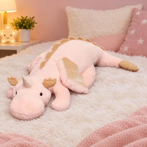 Pode incluir: Um peluche macio em forma de dragão cor-de-rosa com um único chifre na cabeça. A criatura apresenta detalhes em tecido dourado nas asas, no chifre e na crista das costas, com pequenos olhos escuros e um focinho arredondado.