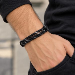 Puede incluir: Una pulsera de cordón negro con un diseño de doble hebra y finas costuras diagonales blancas reflectantes tejidas en el material. La pulsera se asegura con dos nudos deslizantes ajustables en los extremos, creando un aspecto limpio y funcional.