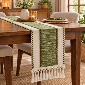Peut inclure: Un chemin de table rectangulaire doté d'un panneau central tissé vert olive, bordé de bandes latérales couleur crème. Le chemin est orné d'une bordure brodée verte complexe et se termine par de longues franges à pompons couleur crème.