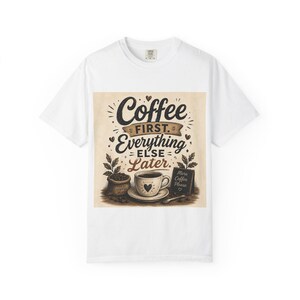May include: A white t-shirt featuring a graphic print with the text Coffee First Everything Else Later. The design includes a coffee cup with a heart, a sack of coffee beans, and a small sign that reads More Coffee Please in brown and black.