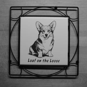 May include: A square white ceramic tile featuring a black and white line drawing of a Corgi dog sitting down. Below the illustration, the text reads Loaf on the Loose. The tile is held within a black metal wire frame with a circular design.