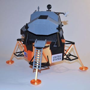 May include: A scale model of a lunar module featuring a gray upper stage and a gold-colored lower stage. The model includes a ladder, landing gear with circular pads, a small United States flag decal, and a sign that reads UNITED STATES.