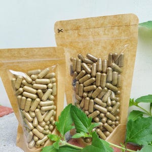 Puede incluir: Dos bolsas verticales de papel kraft marrón claro con ventanas de plástico transparente, llenas de numerosas cápsulas cilíndricas de color verde oliva. Cada bolsa tiene una línea perforada con un icono de tijeras arriba.
