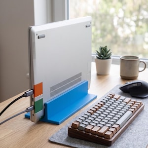 Può includere: Un computer portatile argentato tenuto in posizione verticale da un supporto in plastica blu brillante. Il portatile presenta un piccolo quadrato arancione e uno verde sul lato, con due cavi grigi collegati. Una tastiera meccanica con tasti in legno è posizionata accanto.