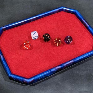 May include: An octagonal dice tray with a blue and purple marbled resin frame and a vibrant red felt interior lining. Four six-sided dice in white, black, orange, and purple are scattered across the red surface within the tray's raised border.