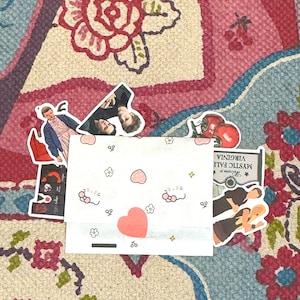 May include: A white envelope decorated with small pink hearts, flowers, and cat faces, containing a collection of stickers. Visible stickers include a person in a blue jacket, two people, red tomatoes, a Mystic Falls Virginia sign, and two figures.
