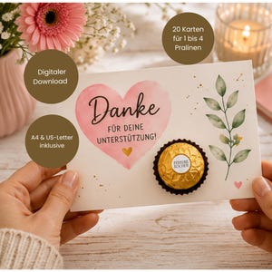 May include: A rectangular card featuring a watercolor pink heart with the German text Danke FÜR DEINE UNTERSTÜTZUNG! and a small gold heart. A single gold-wrapped Ferrero Rocher chocolate is attached to the card, which also displays a green leaf illustration.