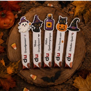 May include: Five rectangular chocolate bars wrapped in white paper with Halloween-themed toppers. The toppers feature a ghost, a black cat in a witch hat, a purple potion bottle, a black cat in a pumpkin, and a witch hat. Each bar has the text du.