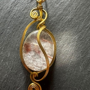 Könnte beinhalten: Ein Anhänger mit einem ovalen, klaren Stein mit roten Einschlüssen. Der Stein ist in einen goldfarbenen Drahtrahmen mit dekorativen Spiralen an der Ober- und Unterseite gefasst und hängt an einer passenden goldfarbenen Kette.