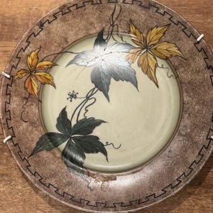 May include: A round ceramic plate featuring a light cream centre with hand-painted dark green, silver, and yellow leaves. The wide rim has a mottled brown finish with a black geometric Greek key border pattern along the inner edge.