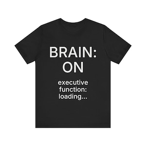 Op de afbeelding: Een zwart t-shirt met korte mouwen en witte tekst op de voorzijde. De tekst luidt: BRAIN: ON executive function: loading... Het t-shirt heeft een ronde hals en een standaard pasvorm, waardoor de opdruk duidelijk zichtbaar is op de stof.