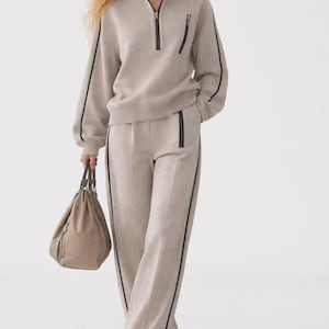 May include: A beige quarter-zip pullover with a black zipper and black side stripes, paired with matching wide-leg trousers featuring black side stripes and a black zippered pocket. A beige drawstring handbag is held at the side.