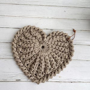 Puede incluir: Un salvamanteles de ganchillo en forma de corazón hecho de cuerda gruesa de color marrón claro. La pieza presenta un patrón de puntos densos y texturizados que irradian desde un orificio central, con una pequeña tira de cuero marrón.