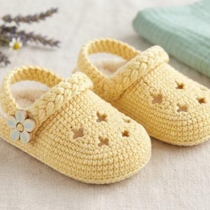 May include: A pair of pale yellow crocheted slippers featuring a braided heel strap, a small white and blue flower-shaped button on the side, and star-shaped cutouts on the top. The interior of the slippers is lined with a soft, cream-colored material.