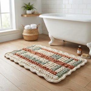 May include: A rectangular crochet bath mat featuring a textured bobble stitch pattern in alternating stripes of cream, sage green, and terracotta. The mat has a scalloped edge border and is laid flat on a light wood floor in a bathroom setting.