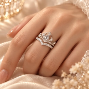 May include: A silver-toned crown-shaped ring set with a central round-cut stone and smaller pavé-set stones along the band. The ring features an intricate filigree design and is paired with a matching V-shaped band adorned with small clear stones.