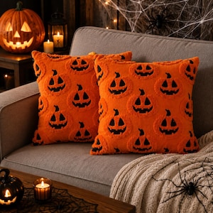 Puede incluir: Dos cojines cuadrados de color naranja vibrante con un patrón repetitivo de caras de calabaza negras. Los cojines tienen una superficie texturizada y con mechones, y están colocados uno al lado del otro sobre un sofá gris tapizado.