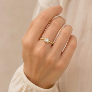Puede incluir: Un anillo de oro con una piedra central rectangular de corte baguette rodeada por un halo de pequeñas piedras redondas. La banda está elaborada con un diseño texturizado de cuentas que se extiende a lo largo de los lados, creando un acabado.