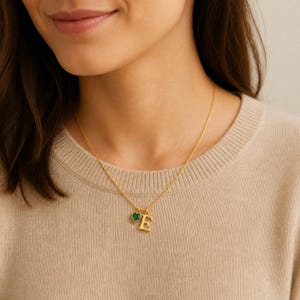 Puede incluir: Un collar de cadena en tono dorado con un colgante de la letra mayúscula E y un pequeño dije en forma de corazón con una piedra verde oscuro. El collar descansa sobre un suéter de punto beige, mostrando un diseño de eslabones delicados.