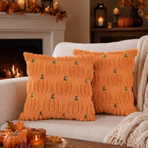 Puede incluir: Dos cojines cuadrados con un patrón repetitivo de calabazas con relieve en color naranja y pequeños tallos verdes. Los cojines tienen una superficie texturizada y están colocados uno al lado del otro sobre un sofá de color claro con un diseño uniforme.