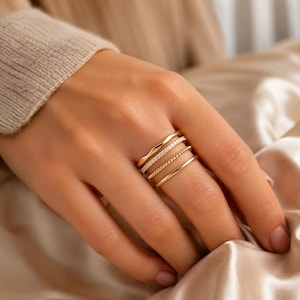 Puede incluir: Un conjunto de cuatro anillos dorados en un dedo. El apilado incluye una banda ondulada lisa, una banda incrustada con pequeñas piedras claras, una banda de cuentas delicada y una banda de oro pulido simple, todas con un acabado amarillo metálico.