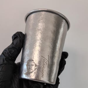 Puede incluir: Un vaso de color plata metálico con un acabado texturizado. El exterior presenta un retrato grabado con láser de una persona con gorra de béisbol, junto a la palabra Titanium escrita en una fuente cursiva en el lateral.