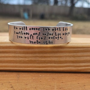 May include: A silver metal bracelet with the words "He will cover you with his feathers, and under his wings you will find refuge. Psalm 91:4" engraved on it.