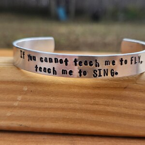 May include: Silver bracelet with the inscription "If you cannot teach me to fly, teach me to sing."