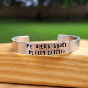 May include: A silver metal bracelet with the text "The Opera Ghost Really Existed" engraved on it.