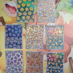 May include: A collection of eight rectangular trading cards featuring repeating character patterns. The cards display Psyduck, Ditto, Pikachu, and Eevee in various colors and holographic finishes, each with printed text including names and stats.