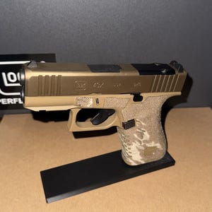 Puede incluir: Una pistola semiautomática con corredera de color bronce y armazón color canela. La corredera tiene grabada la inscripción Glock 43X USA 9x19. La empuñadura presenta un patrón de camuflaje texturizado en tonos canela y marrón sobre soporte negro.