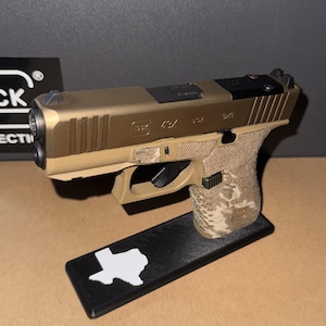 Puede incluir: Una pistola semiautomática modelo 43X de color tostado y negro con empuñadura texturizada, montada sobre un soporte de exhibición rectangular negro con una silueta blanca del estado de Texas. La corredera tiene la inscripción Glock 43X USA 9x19.