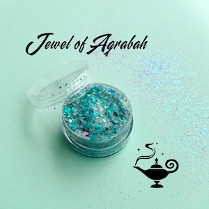 May include: A small, clear, round plastic jar filled with chunky teal, blue, and silver glitter. The lid is open and resting beside the jar. The text "Jewel of Agrabah" is printed above, and a black silhouette of a magic lamp is below.