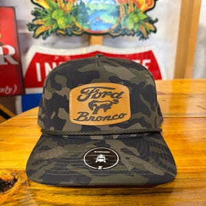 May include: A camouflage print baseball cap featuring a tan leather-like patch on the front with the text Ford Bronco and a bucking horse logo. A dark green braided cord runs across the base of the crown, and a circular Blackhawk sticker is on the brim.