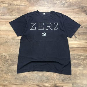 May include: A black short-sleeve t-shirt featuring the word ZERO printed in white capital letters across the chest. Below the text is a small white six-pointed star symbol with a circular center. The shirt has a crew neckline and a standard fit.