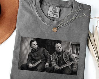 Puede incluir: Una camiseta gris carbón con un estampado gráfico en blanco y negro de Michael Myers y Jason Voorhees, ambos sosteniendo cuchillos. La camiseta tiene cuello redondo, una etiqueta de la marca Comfort Colors y una fina cadena dorada encima.