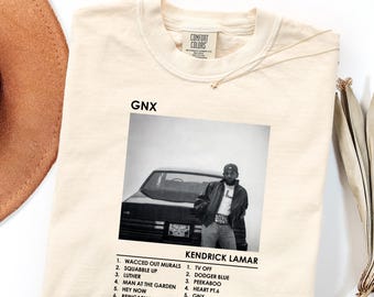 Pode incluir: Uma t-shirt de cor marfim com um gráfico a preto e branco de uma pessoa de pé junto a um carro. O texto GNX e KENDRICK LAMAR aparece acima e abaixo da imagem, com uma lista de doze canções impressa em tinta preta.